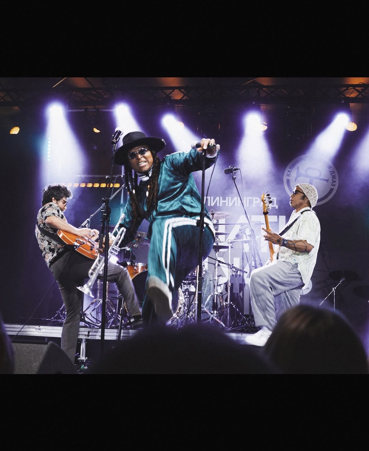 Live band performance with a singer at center stage, guitarist at left, and bassist at right under purple and white lights